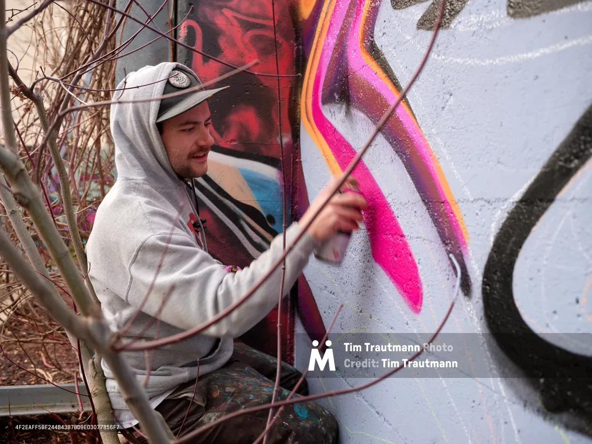 Travis Fields, known as Campographic, concentrates intently as he applies vivid magenta paint to a large-scale mural at the Clay Creative Building in southeast Portland. Framed by bare winter branches, the hooded artist works methodically on the concrete wall, his brush capturing fluid curves against a backdrop of swirling colors and abstract forms. The industrial setting and organic foreground elements create a compelling contrast between urban creativity and natural dormancy.