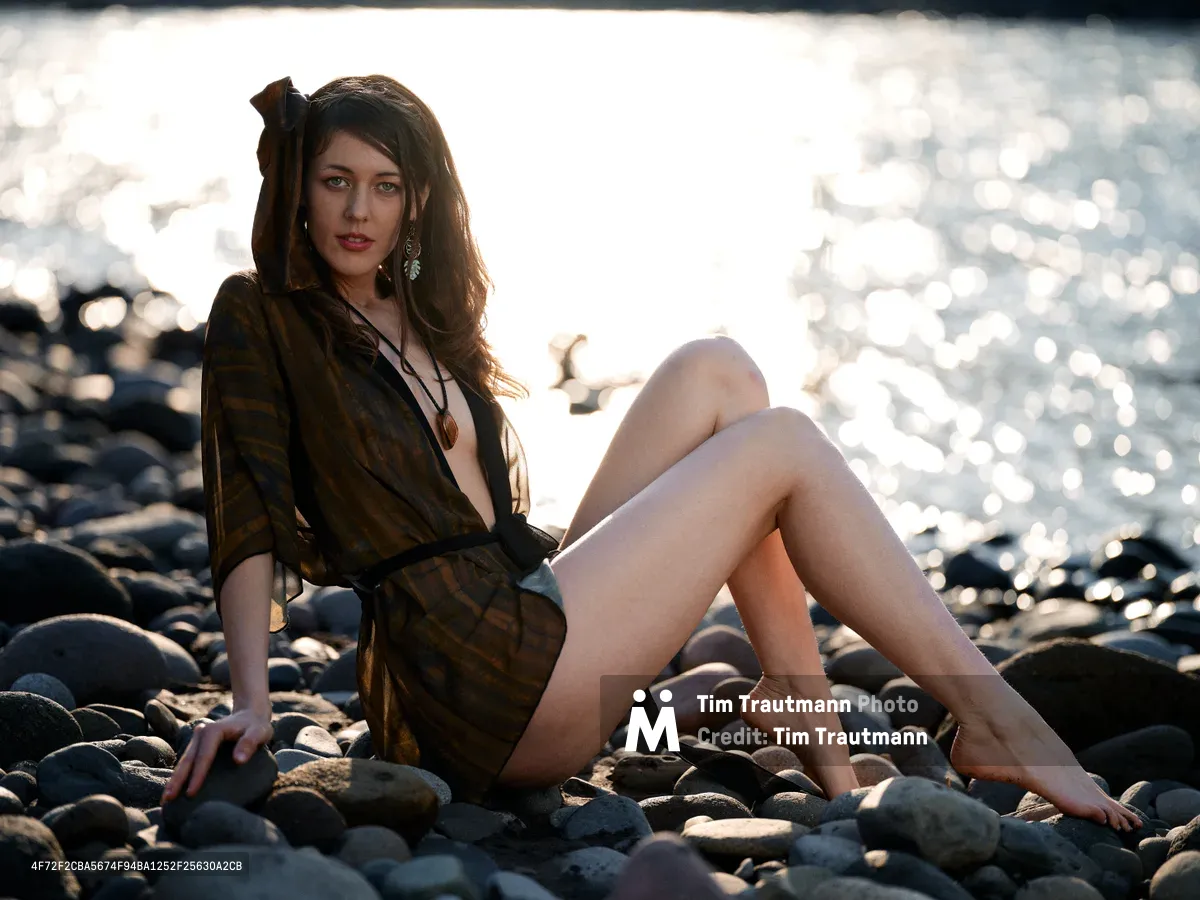 A young woman in a flowing olive wrap dress sits gracefully on the rocky riverbed of the Columbia River near the Floodplain Trail in Multnomah County, Oregon. The golden hour light creates dramatic backlighting as she poses among the smooth river stones, with the sparkling water creating a dreamy bokeh effect in the background. Her long brunette hair catches the warm light while delicate earrings add an elegant touch to the serene riverside portrait. The composition captures both the natural beauty of the Pacific Northwest landscape and an intimate moment of quiet contemplation.