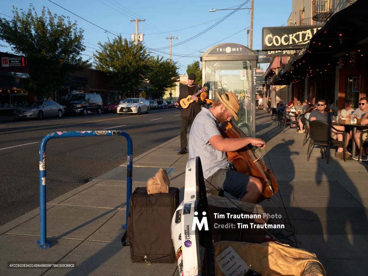Golden hour sunlight illuminates two street musicians performing on Southeast Hawthorne Boulevard in Portland's Richmond neighborhood. A cellist in a straw hat sits prominently in the foreground beside his weathered instrument case, while a guitarist stands behind him near a TriMet bus shelter. The warm evening light casts long shadows across the sidewalk as diners enjoy the outdoor patio atmosphere of nearby restaurants under string lights.