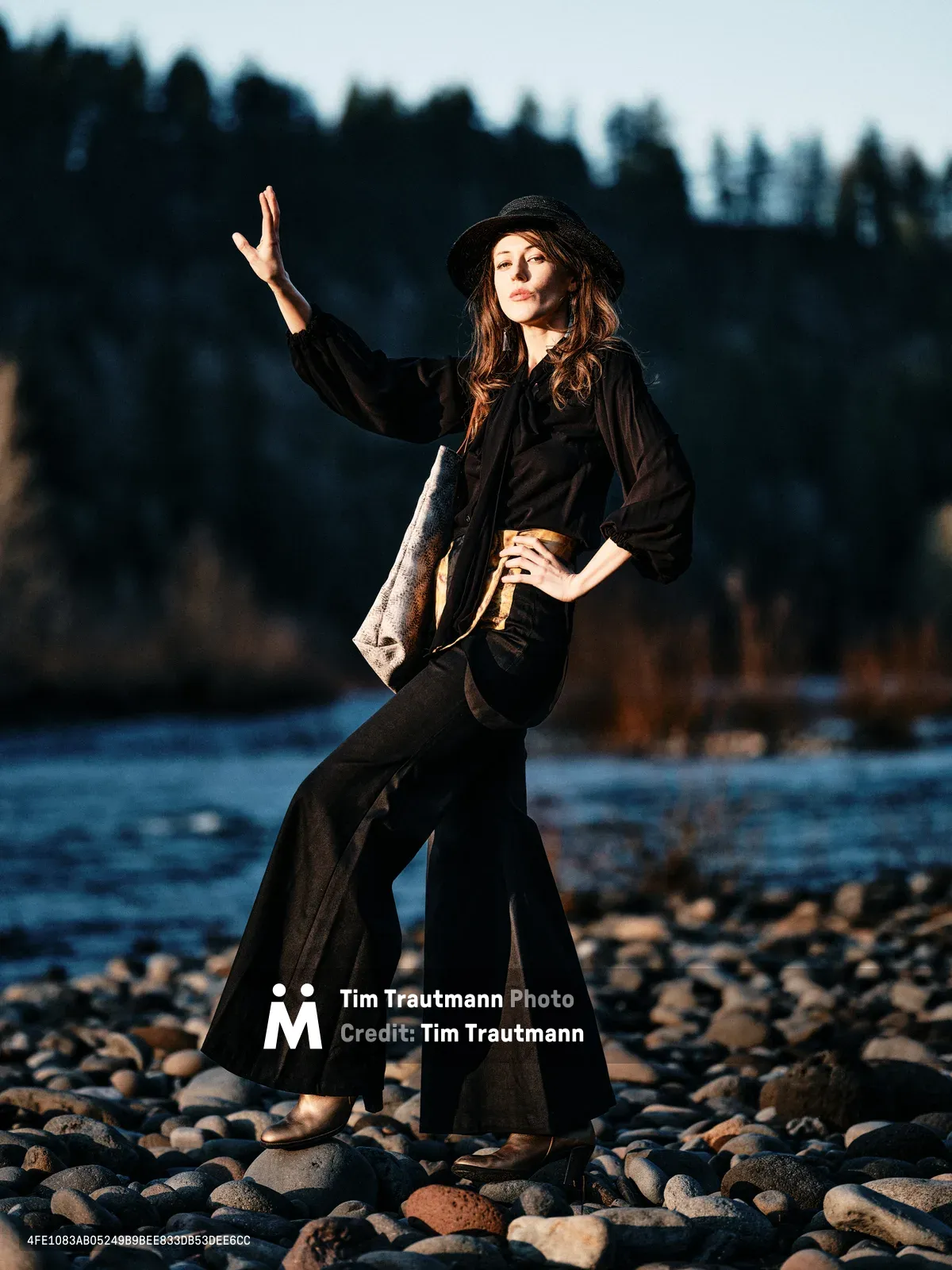 A young woman in flowing black attire and a wide-brimmed hat strikes a theatrical pose on the rocky riverbed of an Oregon waterway, one hand raised toward the sky. Warm golden hour light bathes her figure against the soft blue tones of water and distant forested hills. Her dramatic stance and bohemian styling create an artistic contrast against the natural Pacific Northwest landscape of smooth river stones and evergreen silhouettes.