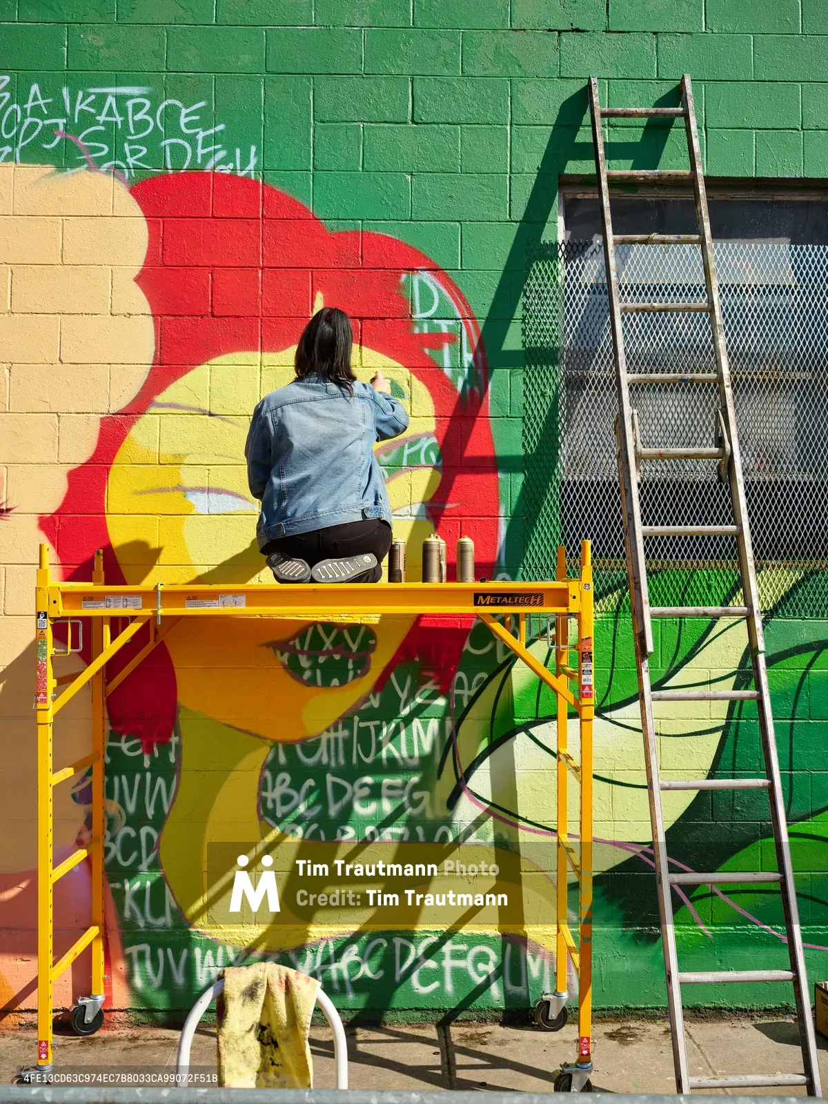 Multidisciplinary artist Vilatose works methodically from a yellow scaffolding platform, adding intricate details to Jardín Cubana, a bold red and green mural depicting three female figures on Northeast Alberta Street. The artist, wearing a denim jacket and dark pants, is captured mid-creation against the geometric brick canvas that transforms this Portland wall into living art. Bright afternoon sunlight casts dramatic shadows across the workspace, while spray paint cans and an aluminum ladder stand ready nearby, testament to the urban artistry flourishing in this historically significant cultural corridor.