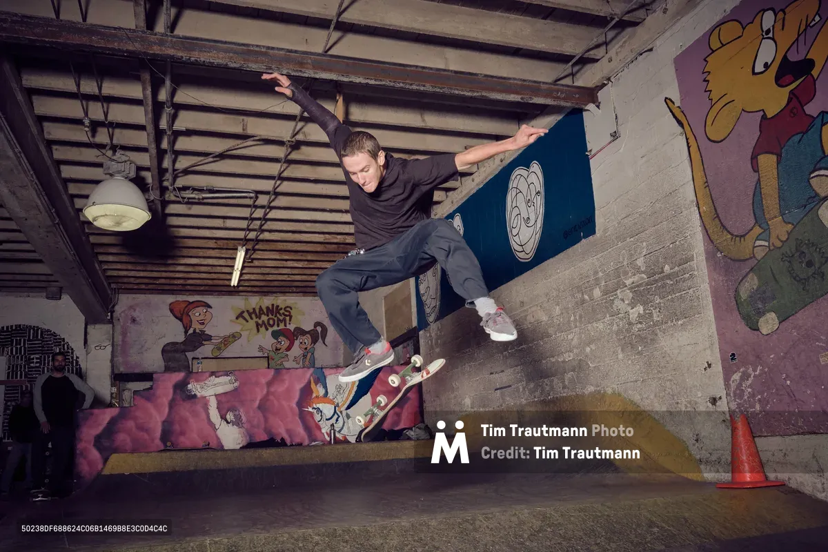 A skateboarder executes a dynamic frontside grind along a concrete ledge in the underground interior of Commonwealth Skateboarding in Portland's Buckman neighborhood. The industrial space features exposed ceiling beams, pendant lighting, and vibrant cartoon murals that create a gritty yet playful atmosphere. The skater's extended arms and focused expression capture the precision required for the technical maneuver, while colorful graffiti art transforms the raw concrete walls into a canvas of urban creativity.