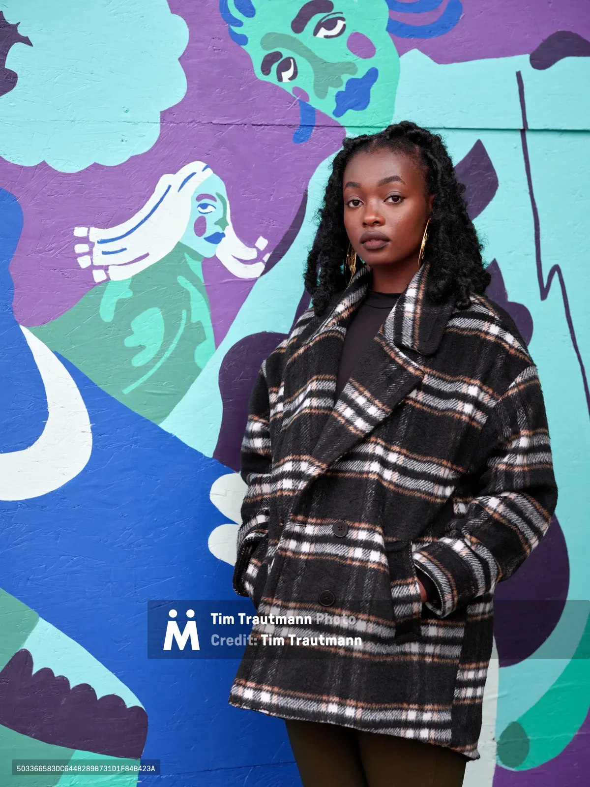 Artist Naomi Likayi stands confidently before her kaleidoscopic mural at Portland's boarded World Trade Center, her plaid wool coat creating striking contrast against the swirling blues, purples, and mint greens of her street art. The composition captures the intersection of personal identity and public art, with Likayi's poised expression reflecting the creative energy radiating from the wall behind her. Commissioned by the Portland Street Art Alliance, the mural transforms urban vacancy into vibrant community expression.