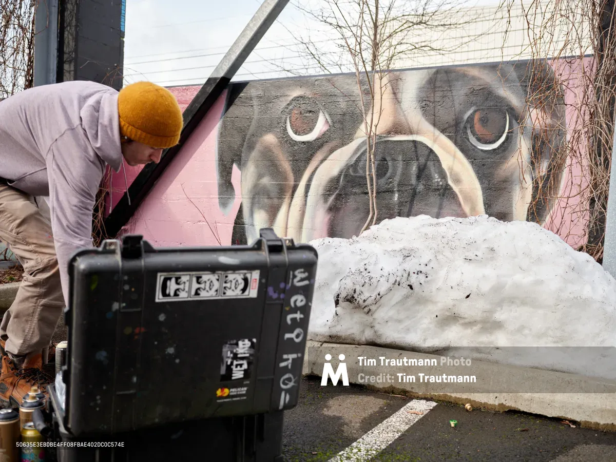 Muralist Wetiko works intently on a striking dog portrait painted on a concrete wall in Portland's Hosford-Abernethy neighborhood. The artist, wearing a golden beanie and light hoodie, leans forward while painting the expressive canine eyes that dominate the pink and gray composition. A film camera on a tripod documents the creative process, capturing both the intimate concentration of the artist and the emerging artwork against the urban backdrop of bare winter branches.