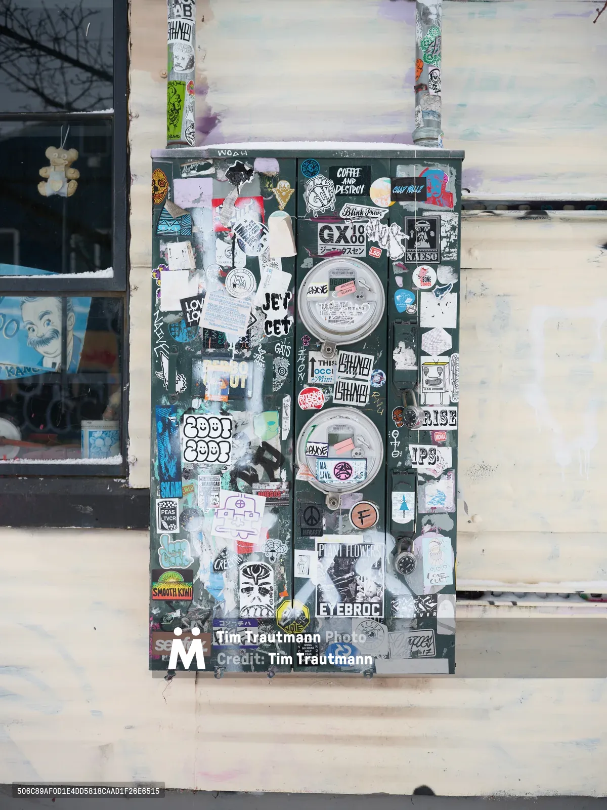 A weathered electrical utility meter on Northeast Alberta Street becomes an impromptu gallery, its dark green surface completely obscured by dozens of street art stickers creating a dense urban collage. The eclectic mix includes hand-drawn characters, band names, political messages, and underground art collective tags layered in organic chaos. Two circular meter faces peek through the accumulated adhesive artwork, while the surrounding cream-colored wall and storefront window frame this testament to Portland's grassroots creative culture in the Concordia neighborhood.