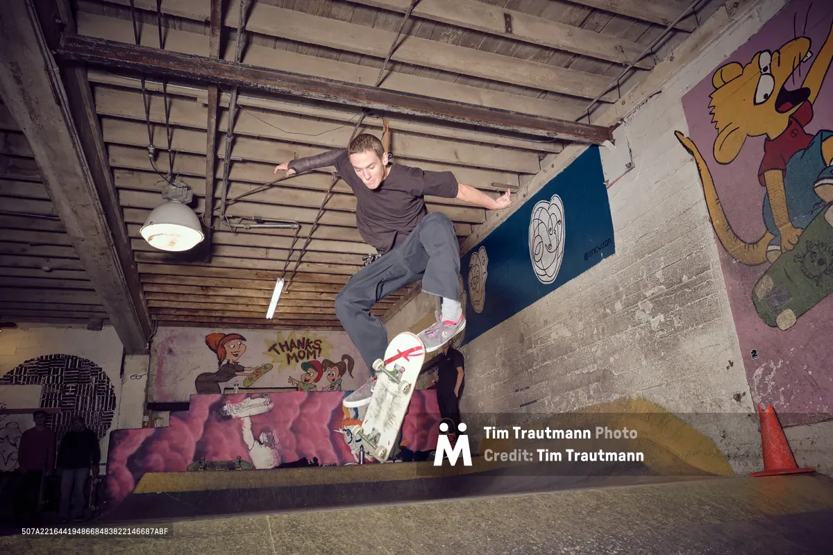 A young skateboarder launches into the air above a concrete bowl at Commonwealth Skateboarding in Portland's Buckman neighborhood, his body twisted in focused concentration. The underground space pulses with creative energy, its exposed ceiling beams and industrial pendant lights casting dramatic shadows across graffiti-covered walls featuring cartoon characters and vibrant murals. The raw concrete transitions create perfect curves for this moment of athletic poetry, captured mid-flight in a space where street culture and artistic expression converge.