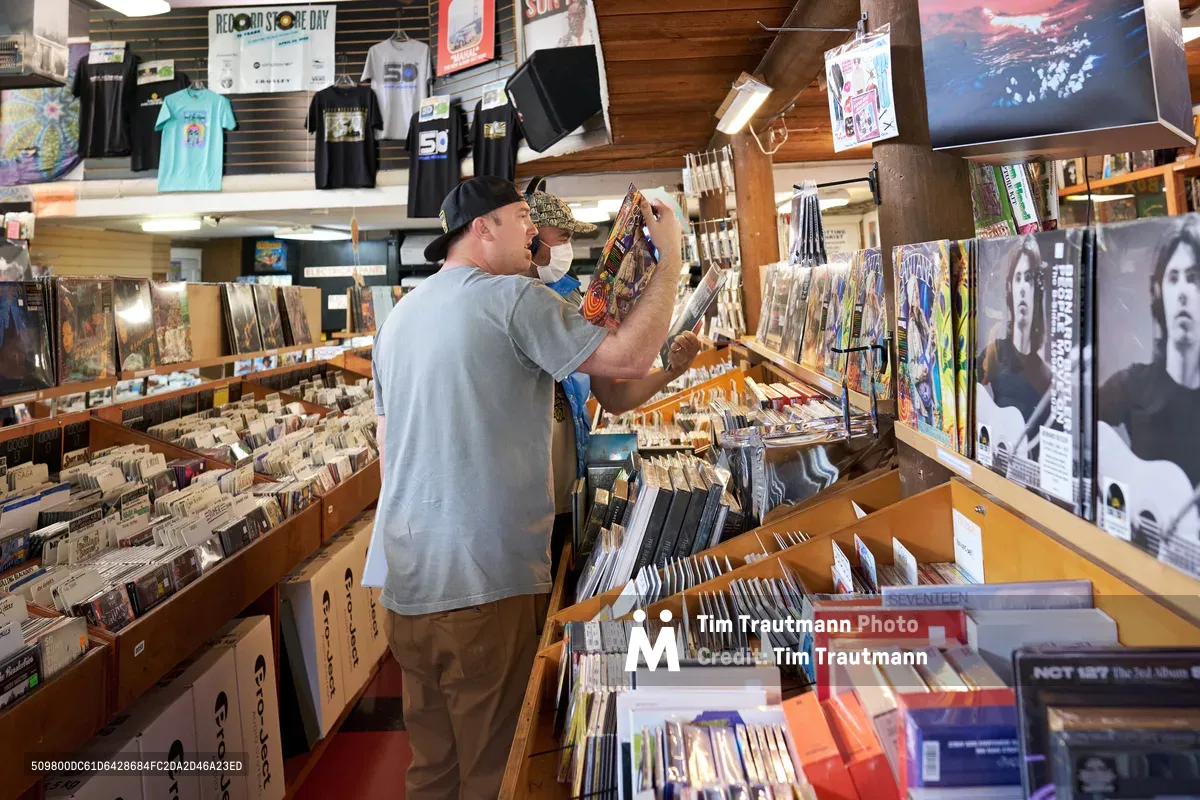 Two customers browse through extensive vinyl collections during Record Store Day 2022 at Music Millennium on East Burnside Street in Portland's Kerns neighborhood. The bearded man in a gray shirt and black cap examines album covers while his companion searches nearby bins. Warm fluorescent lighting illuminates towering wooden shelves packed with records, vintage posters, and band merchandise, creating an intimate atmosphere of musical discovery in this iconic independent record shop.