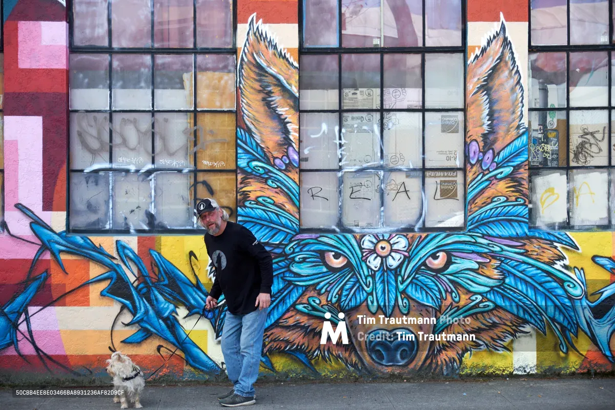 A middle-aged man in casual black attire stands confidently beside his small white terrier against a towering street art mural in Portland's Central Eastside. The vibrant artwork depicts a mystical winged wolf with piercing eyes and azure plumage, its protective gaze watching over the industrial neighborhood. Behind the mural, weathered warehouse windows reflect the soft overcast light typical of the Pacific Northwest, creating an atmospheric backdrop that bridges urban grit with artistic transformation.