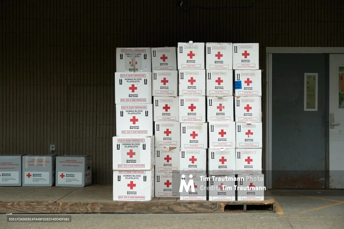Pristine white boxes marked with the iconic red cross emblem stand in orderly formation against weathered corrugated metal siding at the American Red Cross Cascades Region facility in Portland's Eliot neighborhood. Each container bears labels for human blood platelets and temperature requirements, creating a geometric pattern of medical precision against the industrial backdrop. The scene captures the quiet anticipation before these vessels fulfill their vital mission of blood collection and distribution.