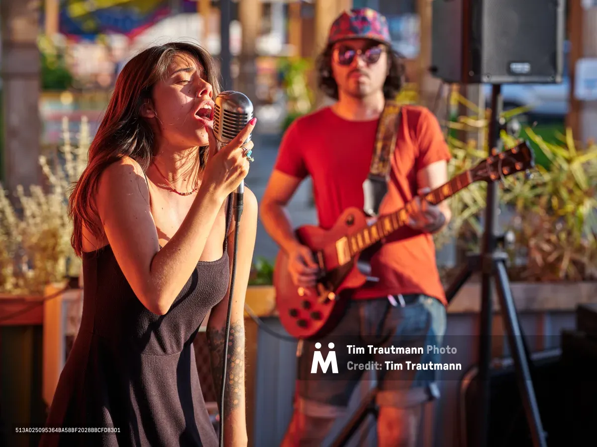 Singer Aubrey Gordon commands the vintage microphone in golden afternoon light, her auburn hair catching the warm glow as she performs with passionate intensity. Behind her, a guitarist in red shirt and patterned cap provides melodic accompaniment, their duo framed by the vibrant outdoor marketplace atmosphere of Portland Mercado. The rich amber light creates dramatic shadows and highlights, transforming this Southeast Portland venue into an intimate concert stage. Lush greenery and colorful market stalls blur into an atmospheric backdrop, emphasizing the raw energy of live street performance.