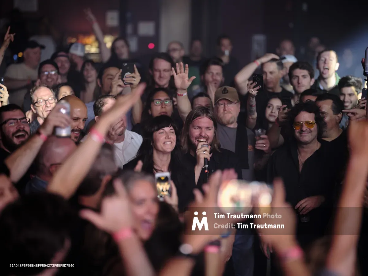 Band of Skulls members Matt Hayward and Emma Richardson perform in the middle of a packed, energetic crowd at Webster Hall in New York City in September 2019. Matt, with long hair and wearing a black shirt, holds a microphone and smiles broadly, while Emma stands beside him. Audience members surrounding them cheer, raise their hands, and hold up smartphones to capture the moment. The dimly lit venue is filled with excitement and motion blur in the foreground.