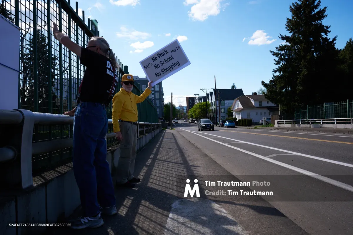 Two activists stand on North Skidmore Street in Portland's Humboldt neighborhood, holding a protest sign calling for a May 1st General Strike with the message "Stop No Work No School No Shopping." The demonstrators are positioned along Interstate 5, with one wearing a bright yellow jacket and cap while the other sports denim overalls. Late afternoon sunlight casts long shadows across the sidewalk as fluffy white clouds drift through a brilliant blue sky above the urban landscape of mixed residential and modern glass buildings.