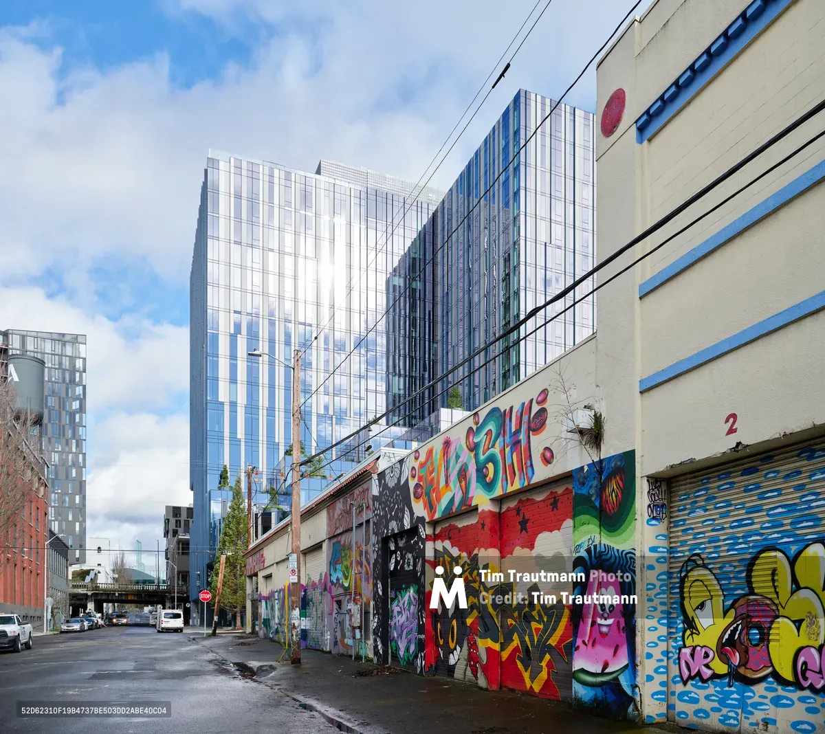 Vibrant street art transforms a weathered industrial wall in Southeast Portland's Central Eastside district, where commissioned murals by the Portland Street Art Alliance breathe life into the former Salvation Army warehouse. The graffiti-style artwork erupts in bold yellows, pinks, blues, and reds against cream-colored concrete, while gleaming glass towers of the city's modern development loom dramatically overhead. Power lines slice across a partly cloudy sky, creating a striking juxtaposition between grassroots artistic expression and corporate architecture.