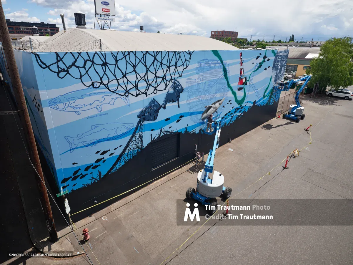 Artists Jeremy Nichols and Devin Finley work from blue scissor lifts to complete an expansive aquatic-themed mural on a warehouse wall in Southeast Portland's Central Eastside district. The sweeping composition features fishing nets, marine life, and nautical elements rendered in blues and teals across the industrial building's facade. Orange traffic cones and safety equipment dot the concrete staging area below, while the artists methodically bring their oceanic vision to life under partly cloudy skies.