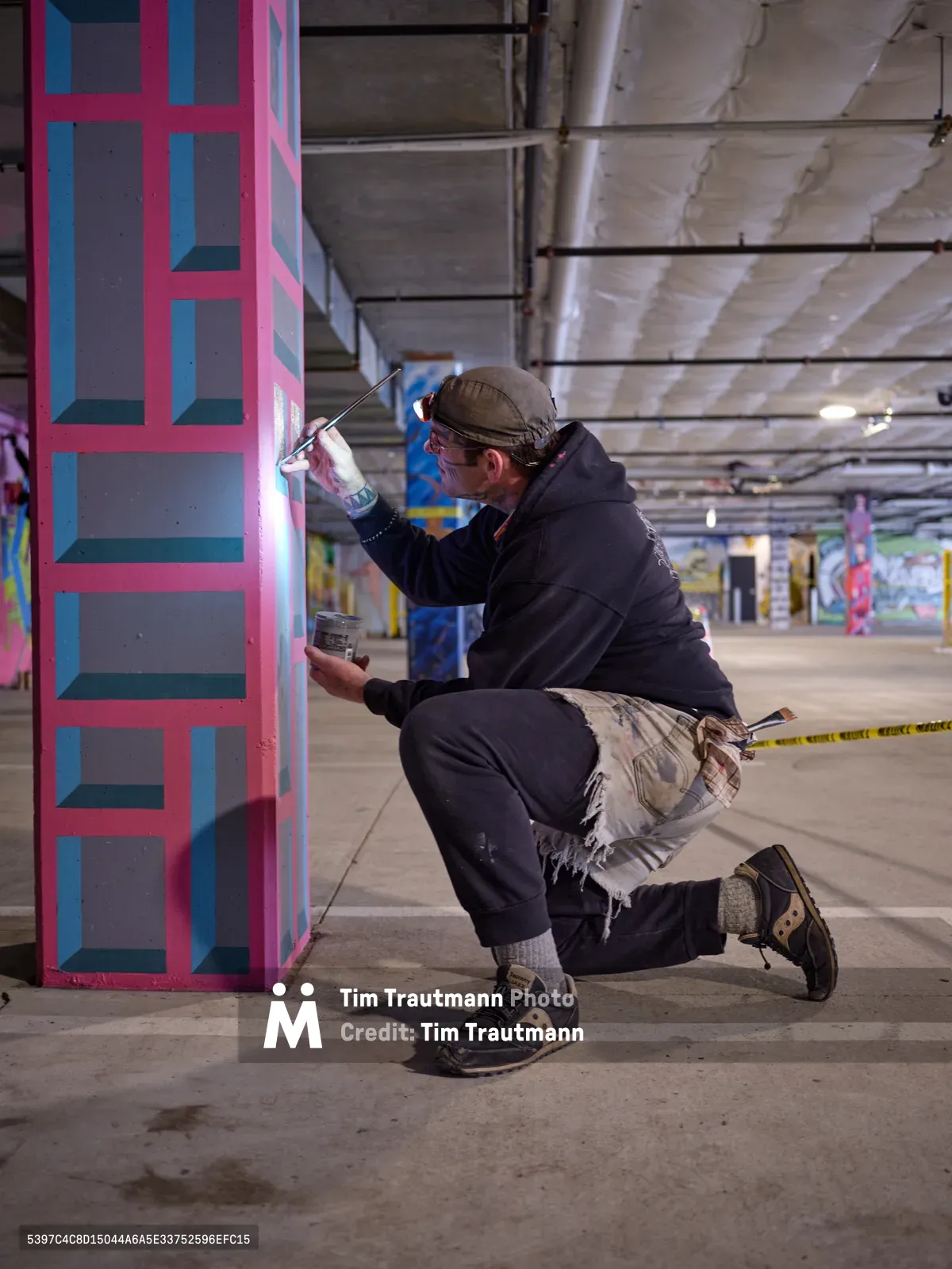 Portland bike activist and artist Rabbitt kneels in concentrated focus as he applies spray paint to a concrete pillar in the Clay Creative building's parking garage. His geometric composition features bold pink and blue rectangular forms that contrast sharply against the raw industrial architecture. The underground space, illuminated by harsh fluorescent lighting, transforms into an impromptu studio where urban art meets utilitarian infrastructure.