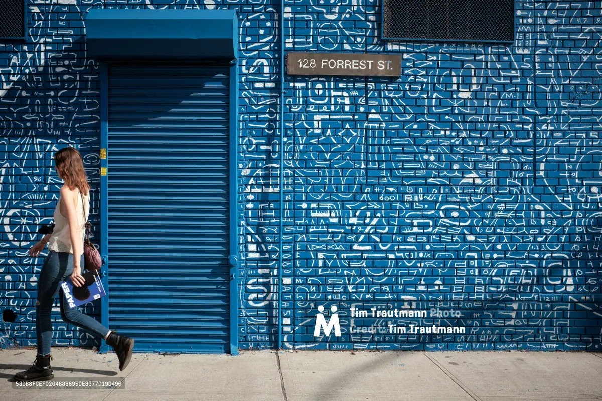 A woman in casual attire strides purposefully past an extraordinary blue mural that transforms the entire facade of 128 Forrest Street in Brooklyn into a mesmerizing maze of white geometric patterns and symbols. The monochromatic artwork creates an optical illusion of depth and movement, with intricate line work covering every surface from the roll-up security gate to the surrounding walls. The pedestrian's natural movement provides a striking human counterpoint to the static yet visually dynamic street art, while bright daylight enhances the vivid cobalt blue that dominates the urban canvas.