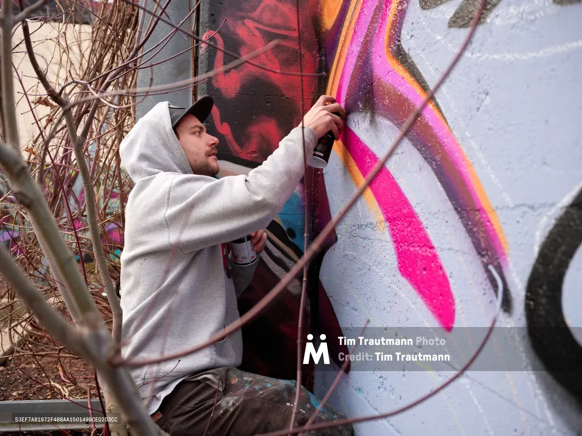 Travis Fields, known as Campographic, meticulously applies vibrant magenta and orange spray paint to a concrete wall at the Clay Creative Building in Portland's Hosford-Abernethy neighborhood. Captured mid-creation, the hooded artist works with focused intensity among dormant winter branches, transforming the urban canvas with bold geometric patterns. The industrial setting frames this moment of artistic genesis, where raw concrete surrenders to explosive color.