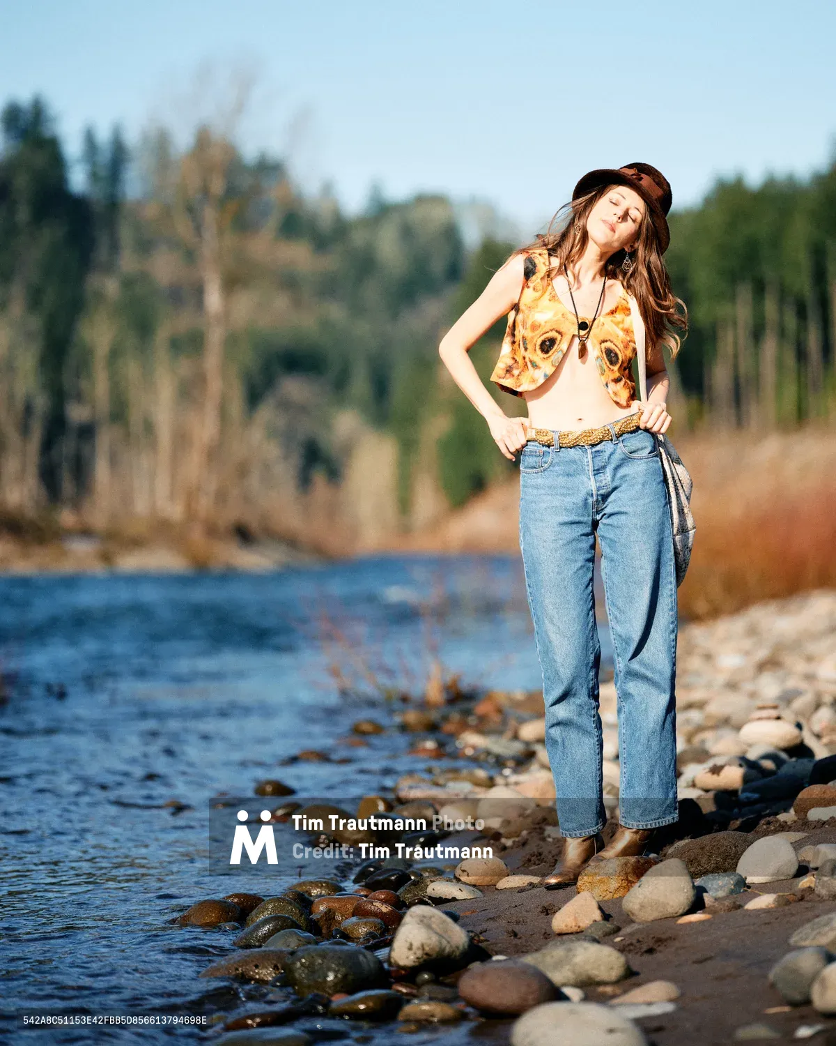 A young woman with flowing brown hair tilts her head back in peaceful contemplation, standing barefoot on the rocky riverbed of Oregon's Floodplain Trail. Bathed in warm golden light, she wears a fox-print crop top, high-waisted jeans, and a dark fedora, her eyes closed in serene meditation. The Columbia River's gentle waters lap against smooth river stones while autumn trees blur softly in the background, creating an intimate moment of connection with the Pacific Northwest wilderness.