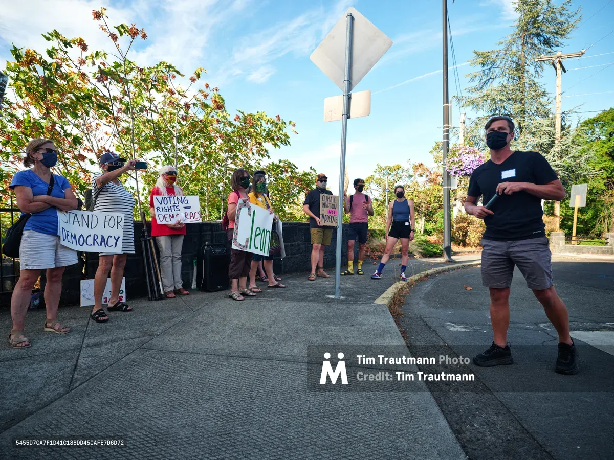 Under a crystalline autumn sky in Oregon City, organizer Michael Gaskill addresses a gathering of masked activists assembled outside Representative Kurt Schrader's McLoughlin Promenade office. The diverse group of protesters holds handwritten signs demanding voting rights and democratic accountability, their casual summer attire and face coverings capturing the intersection of pandemic-era activism and civic engagement. Golden afternoon light filters through the leafy canopy overhead, casting dappled shadows across the concrete sidewalk where democracy advocates make their voices heard.