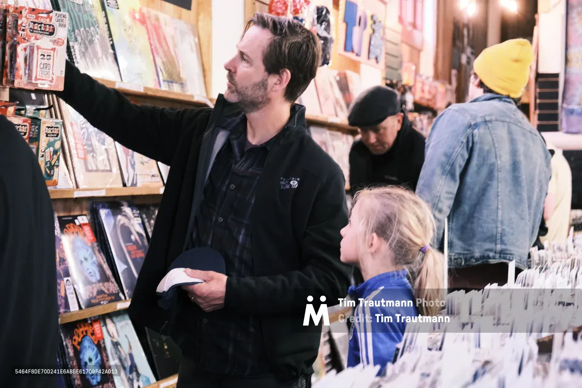A bearded father in a dark jacket reaches up to browse vinyl records on the upper shelves while his young blonde daughter in a blue tracksuit watches from below at Portland's iconic Music Millennium record store during Record Store Day. The warm, intimate scene captures the intergenerational sharing of music discovery amid towering walls of albums and the soft glow of the shop's ambient lighting. Other customers browse quietly in the background, creating an authentic atmosphere of musical exploration on this celebrated day for independent record stores.