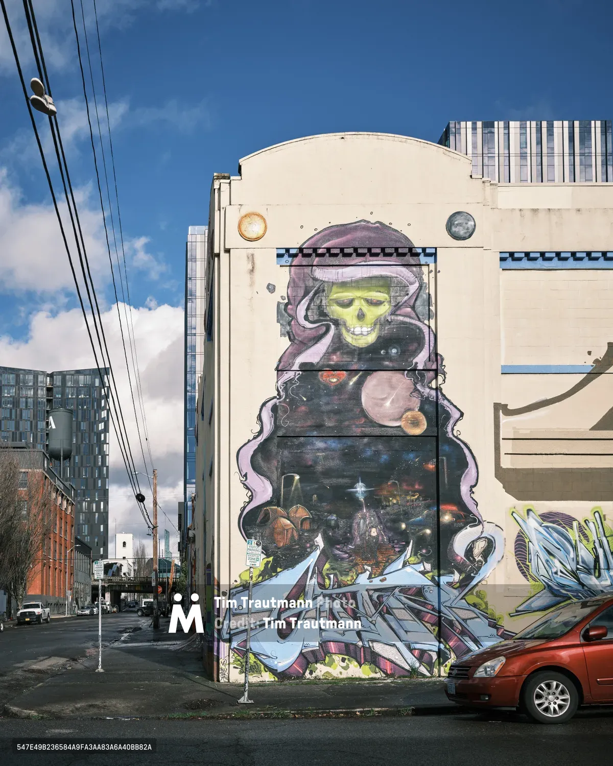 A towering street mural transforms the curved facade of a former Salvation Army warehouse on Southeast Ash Street in Portland's Central Eastside district. The artwork depicts a haunting lime-green skull wreathed in purple cosmic smoke, its hollow sockets gazing over a star-filled cosmic landscape populated with planets and celestial debris. Power lines slice diagonally through the bright blue afternoon sky, while modern apartment buildings frame the scene and a red sedan parks casually beneath this monumental display of urban art.