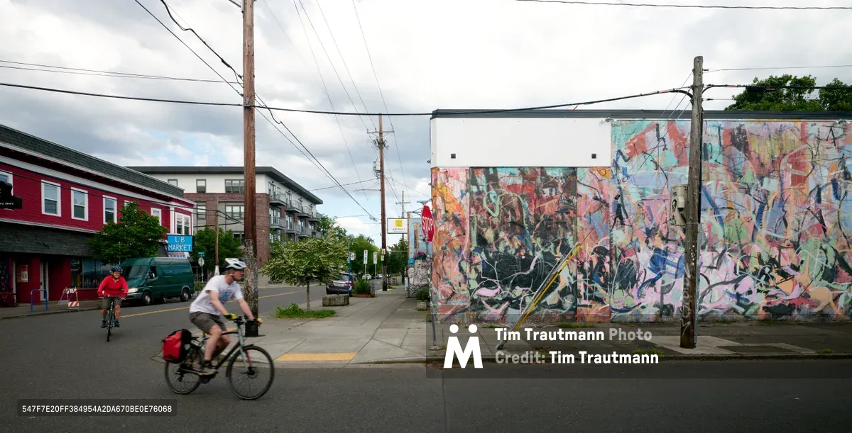 Two cyclists traverse Southeast Division Street in Portland's Richmond neighborhood, gliding past an expansive abstract mural that transforms a weathered building wall into a canvas of vibrant chaos. The overcast afternoon light softens the scene while power lines create geometric counterpoints to the fluid graffiti art. Red residential buildings anchor the left side of the frame, establishing the authentic neighborhood character of this creative corridor.