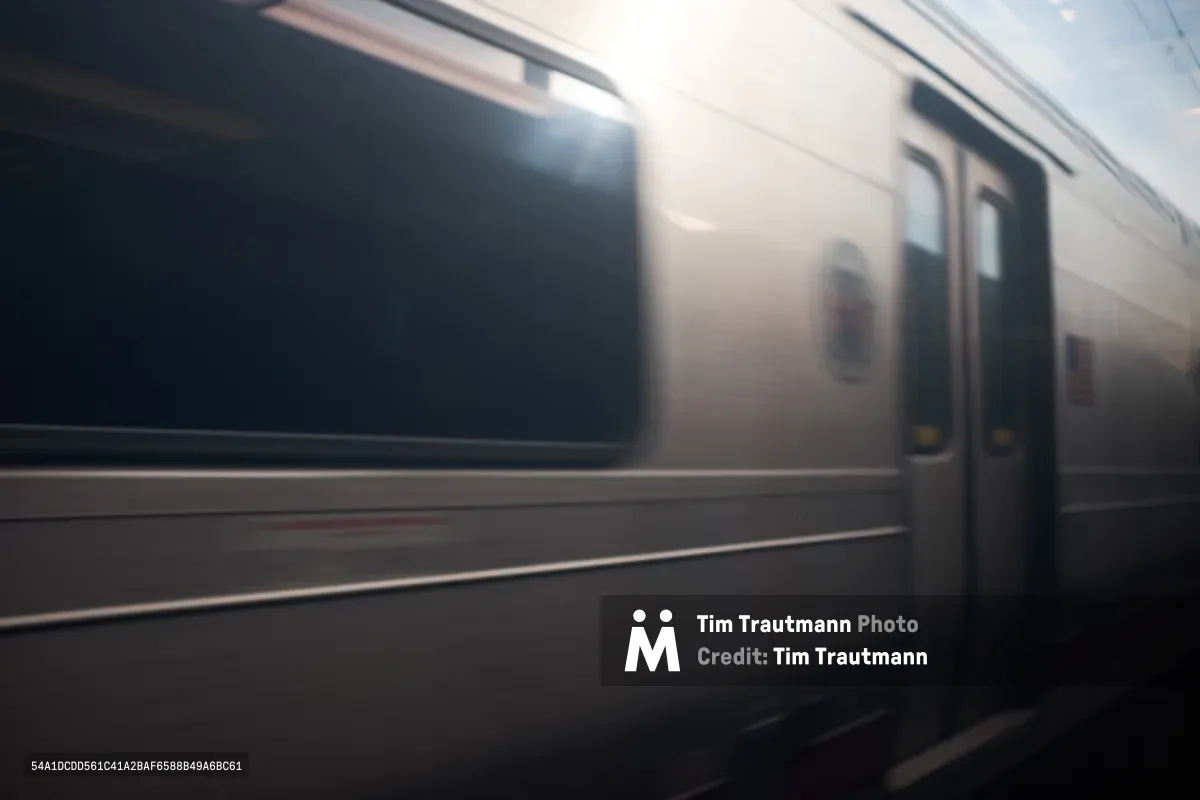 A silver Amtrak passenger train car rushes past the platform at Kearny station in Hudson County, New Jersey. Motion blur captures the speed and energy of the departing train, with the dark platform contrasting against the illuminated car windows. The atmospheric shot emphasizes the fleeting nature of rail travel through the industrial corridor near Newark.