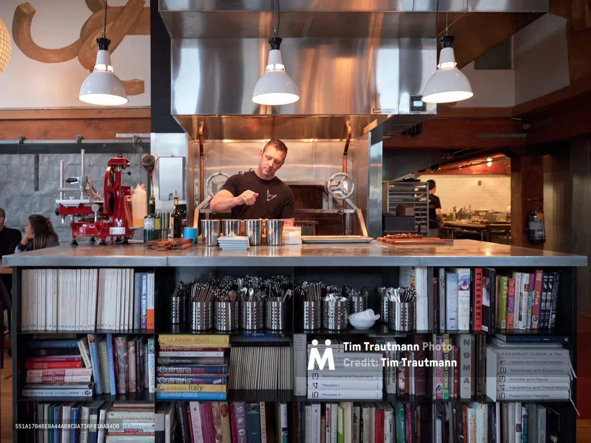 A dedicated chef works intently at a professional kitchen island beneath gleaming copper ventilation hoods in this Portland restaurant. The warm industrial space features exposed wooden beams, pendant lighting, and an extensive cookbook collection housed in built-in shelves beneath the polished steel counter. Steam and golden light create an atmospheric backdrop as the culinary professional focuses on his craft in this unique kitchen-library hybrid at Plaza del Toro in the Buckman neighborhood.