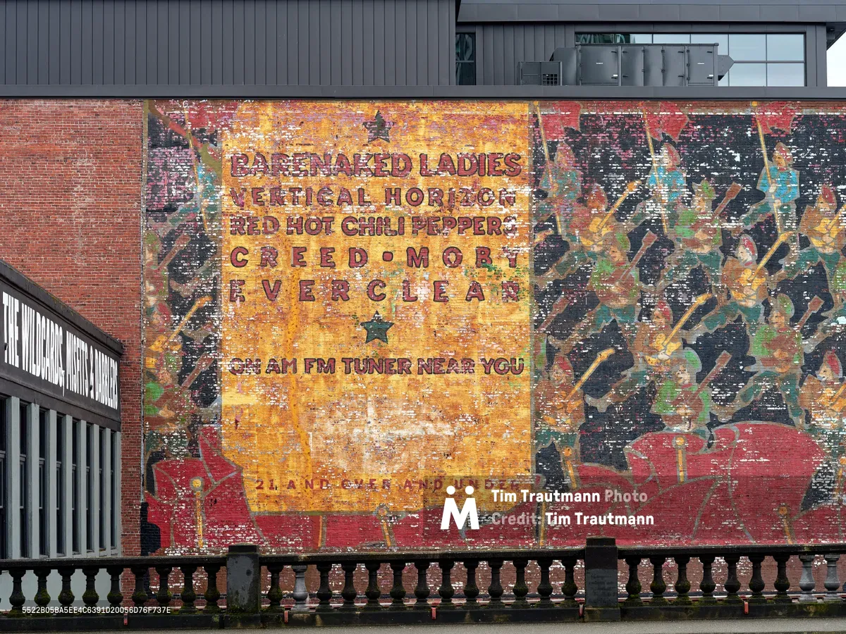 A weathered vintage billboard mural on a brick building near Portland's Burnside Bridge advertises alternative rock station KNRK 94.7 FM, featuring bands like Barenaked Ladies, Vertical Horizon, Red Hot Chili Peppers, Creed, Moby, and Everclear. The painted advertisement shows significant aging and peeling, with its golden yellow center panel contrasting against red and black geometric patterns that frame the radio station branding. Viewed from behind ornate iron railings, this ghost sign captures the ephemeral nature of commercial art in urban landscapes.