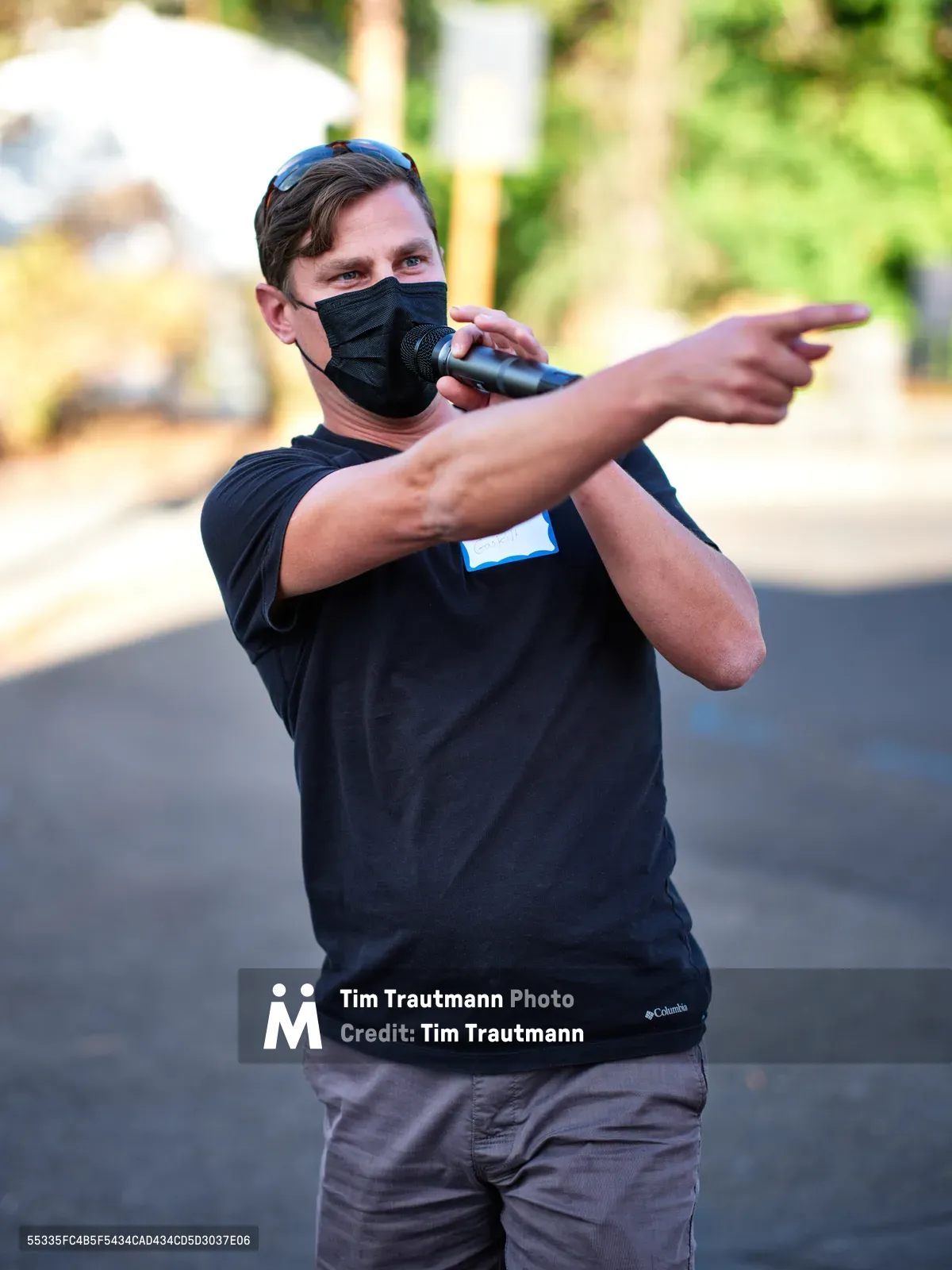 Michael Gaskill, wearing a black face mask and Columbia shirt, gestures emphatically while speaking into a microphone during a demonstration outside Representative Kurt Schrader's Oregon City office. Golden afternoon light filters through the trees behind him, creating a warm bokeh backdrop as he addresses the assembled crowd on McLoughlin Promenade. His animated stance and pointing gesture convey passionate advocacy, captured in this moment of civic engagement.