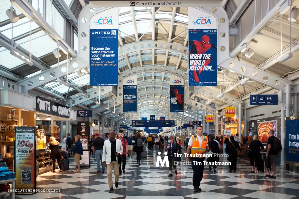 The soaring barrel-vaulted ceiling of Chicago O'Hare International Airport creates a luminous cathedral of transit, where streams of travelers navigate the polished checkerboard floor beneath suspended airline advertisements. Natural light filters through the architectural skylights, illuminating the constant flow of passengers moving between gates while airport personnel in high-visibility vests maintain operational order. The convergence of steel beams and glass creates dramatic perspective lines that draw the eye toward the terminal's vanishing point, where departure gates await.