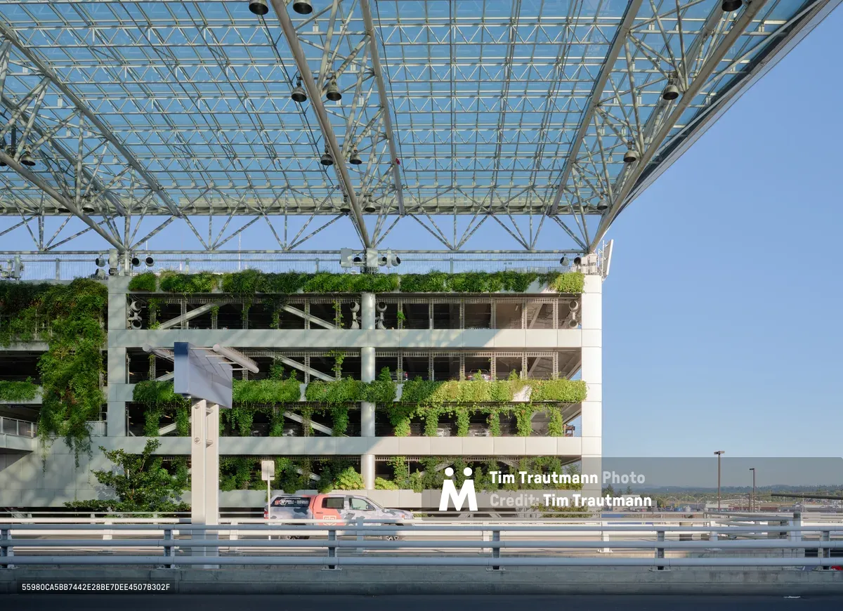 A striking view of Portland International Airport's innovative short-term parking garage showcases the marriage of sustainable design and modern infrastructure. Cascading vegetation transforms the concrete structure into a verdant vertical garden, while an intricate steel and glass canopy overhead creates geometric patterns against the clear Oregon sky. The foreground highway barrier frames this architectural achievement, where a lone red vehicle provides scale to the ambitious green building initiative that defines PDX's commitment to environmental stewardship.