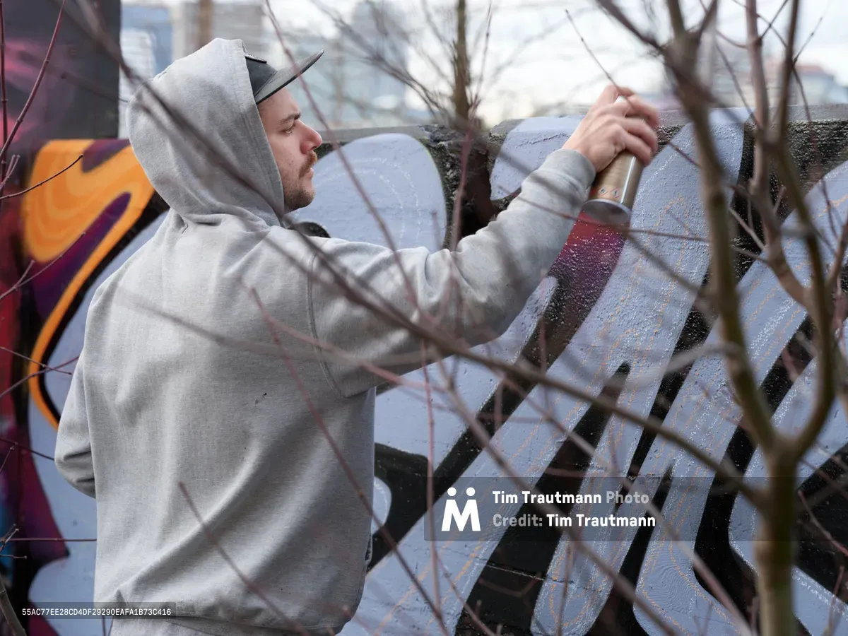 Travis Fields, known as Campographic, concentrates intently as he applies paint to a weathered concrete wall at the Clay Creative Building in southeast Portland's Hosford-Abernethy neighborhood. Clad in a gray hoodie and black cap, the muralist works with deliberate precision while bare winter branches frame the foreground, creating natural geometry against the urban canvas. The soft daylight illuminates his focused expression and the textured surface where vibrant colors begin to emerge against the industrial backdrop.