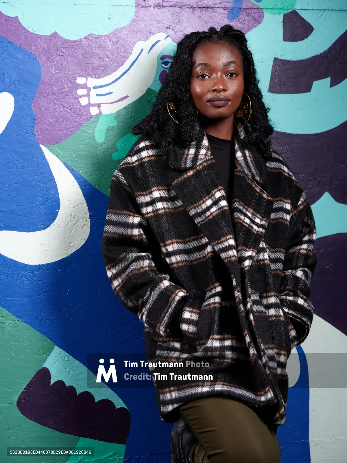 Artist Naomi Likayi stands confidently before her vibrant mural at Portland's boarded World Trade Center, her black protective locs framing her contemplative expression. She wears an oversized plaid coat in earth tones that complements the swirling blues, purples, and greens of her abstract artwork behind her. The portrait captures both the artist's quiet determination and the dynamic energy of her public art commission, creating a powerful dialogue between creator and creation in downtown Portland's urban landscape.