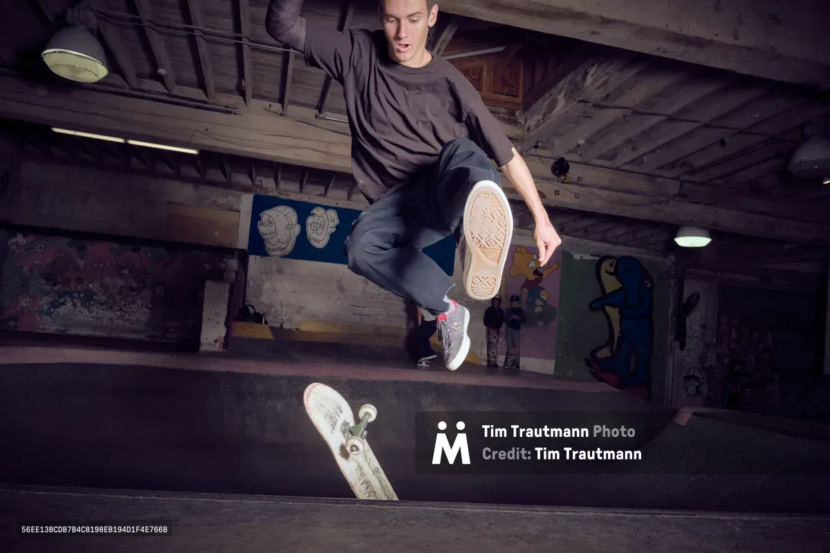 A young skateboarder executes a dynamic trick in the shadowy depths of Commonwealth Skateboarding's concrete bowl in Portland's Buckman neighborhood. The industrial ceiling beams and graffitied walls create a gritty underground atmosphere, while moody lighting illuminates the skater's focused expression mid-maneuver. The worn concrete transitions and scattered deck below speak to countless sessions in this legendary Southeast Portland skate haven.