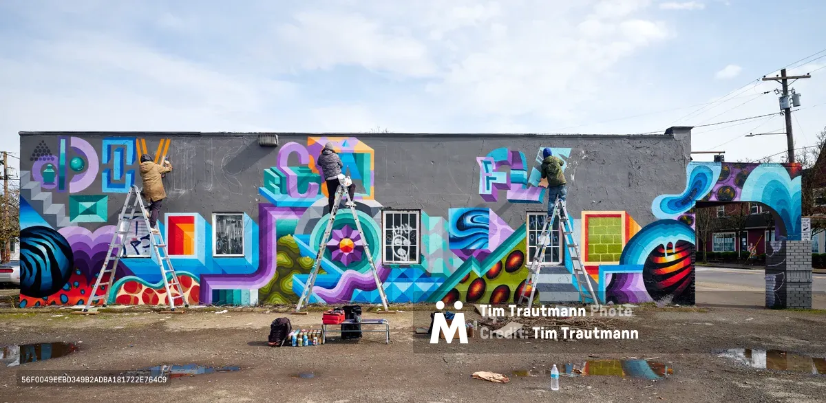 Three artists work simultaneously on an expansive geometric mural covering a gray industrial building in Portland's urban landscape. Perched on aluminum ladders, they paint vibrant interlocking shapes in electric blues, purples, and greens that spell out abstract letterforms across the concrete facade. The overcast Pacific Northwest sky provides soft, even lighting while spray cans, cardboard, and painting supplies scatter across the weathered asphalt below, creating an authentic documentation of street art in progress.