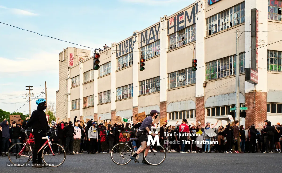 Two cyclists provide traffic direction and crowd protection as hundreds of demonstrators gather along Northeast 33rd Avenue holding Black Lives Matter protest signs in the wake of George Floyd's murder. The late afternoon light bathes the historic industrial buildings while traffic lights punctuate the urban scene. The cyclists serve as guardians of the peaceful assembly, managing the intersection between vehicular traffic and the stationary crowd in Portland's Grant Park neighborhood.