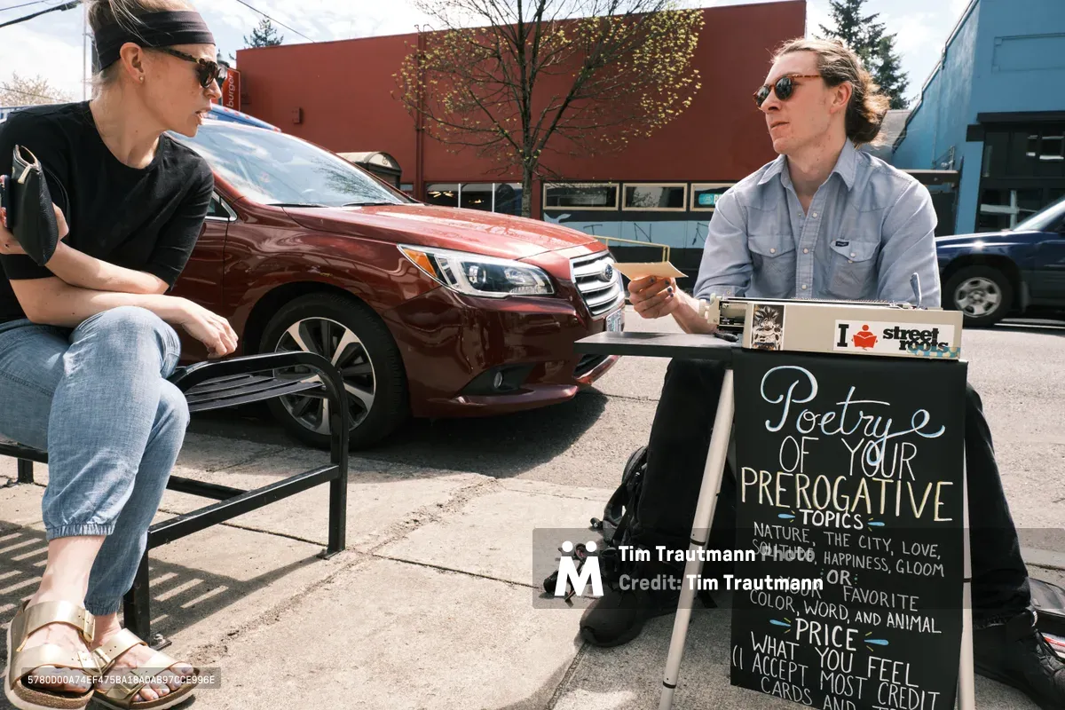 On a sun-drenched afternoon along Northeast Alberta Street in Portland, street poet Till Gwinn crafts personalized verse for a customer seated nearby. The artist works at his portable setup with legs spread apart as he sits at his typewriter, complete with a chalkboard advertising custom poetry on topics ranging from nature to love, while his patron waits patiently beside a burgundy sedan. The scene captures the intimate exchange of art and commerce in Portland's vibrant Alberta Arts District, where creative entrepreneurs transform sidewalks into stages for their craft.