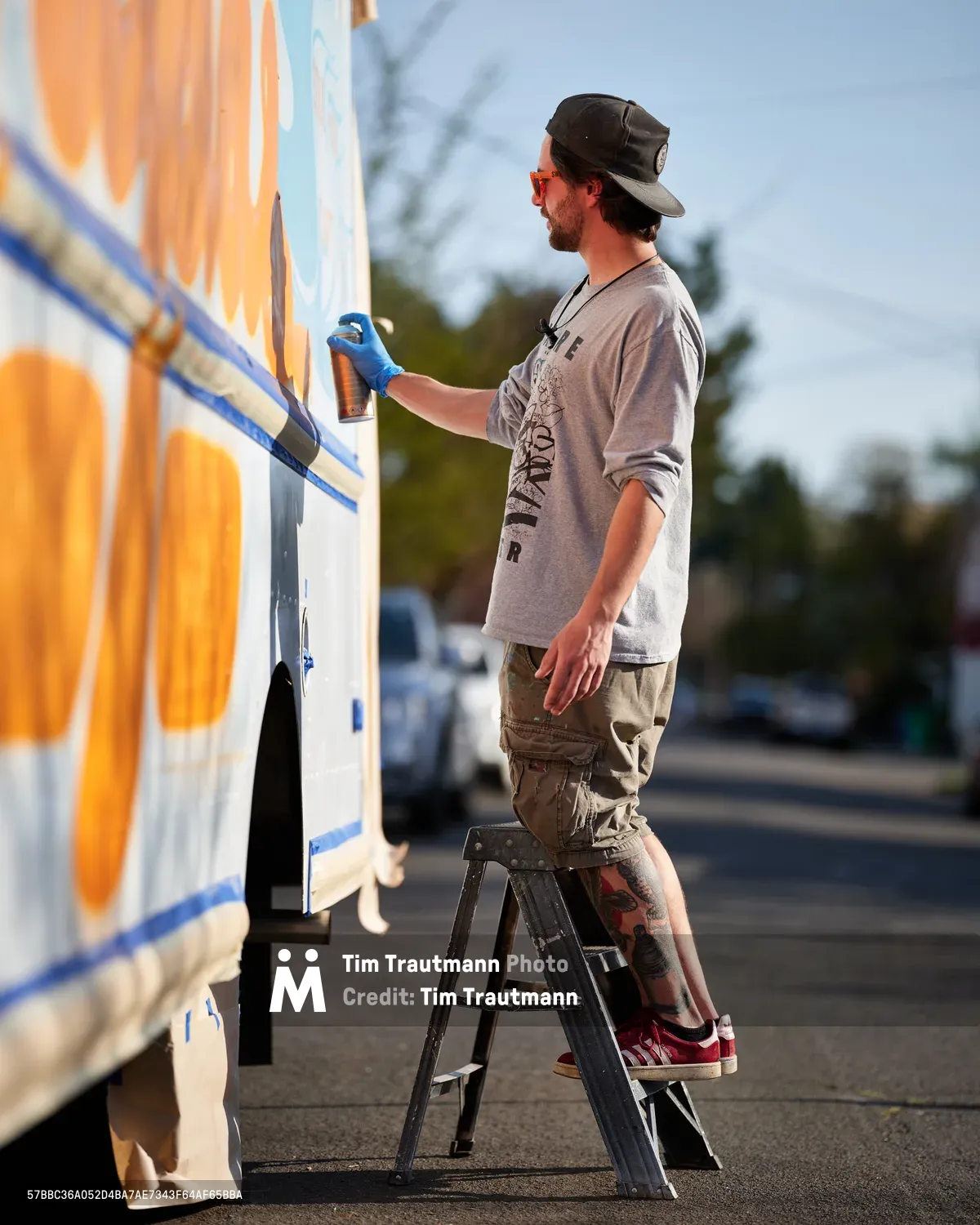 Golden hour light bathes a bearded muralist as he carefully applies spray paint to the side of a white food truck on Northeast 9th Avenue in Portland, Oregon. Standing on a weathered stepladder, the artist wears a black cap and gray long-sleeve shirt while working on vibrant orange and blue geometric designs. The warm evening light creates a luminous contrast against the emerging artwork, capturing the intersection of mobile commerce and street art culture in this King neighborhood scene.