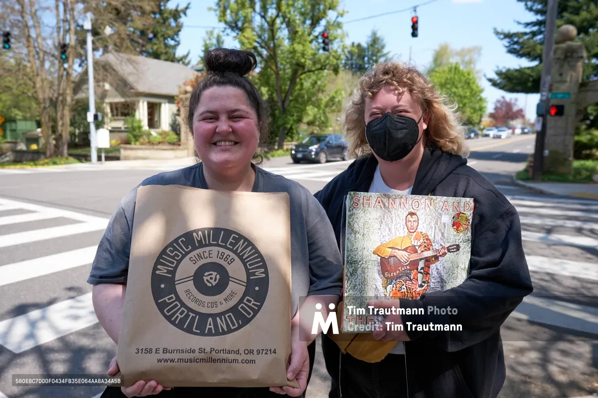 Two friends stand proudly outside Music Millennium on East Burnside Street during Record Store Day 2022, clutching their vinyl discoveries against the backdrop of Portland's leafy Kerns neighborhood. The woman on the left beams with joy while holding a Music Millennium shopping bag, her dark hair twisted into a casual top knot. Her companion, wearing a black mask and hoodie, displays a vintage Shannon and The Clams album cover, their curly blonde hair catching the filtered spring sunlight. Behind them, the quiet residential street stretches toward a traffic light, lined with mature trees and characteristic Portland homes.