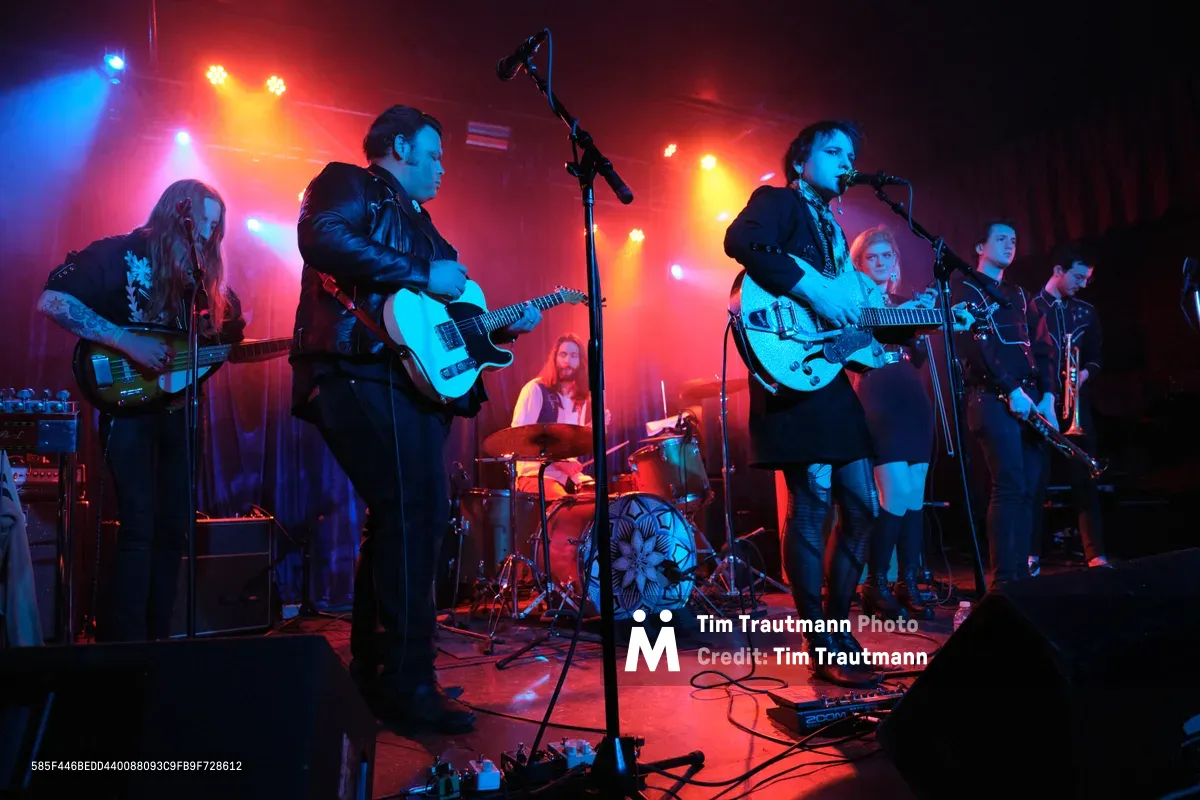 Roselit Bone commands the intimate stage at Dante's in Portland, bathed in crimson and azure stage lights that pierce through the venue's signature darkness. The band's frontwoman grips her white Gretsch guitar while delivering vocals into the microphone, flanked by leather-clad bandmates wielding electric guitars and bass. Behind them, the drummer maintains rhythm beneath a geometric kick drum design, while the warm glow of amber spotlights creates dramatic silhouettes against the club's moody atmosphere.
