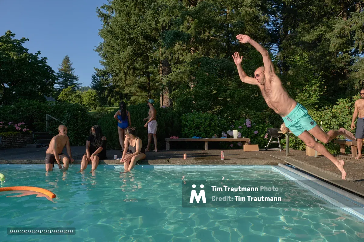 A shirtless man in mint green swim trunks leaps dynamically through the air above a crystalline swimming pool at Orchard Vibration in Newberg, Oregon. His body forms a graceful arc against the backdrop of towering evergreens and azure sky, while friends gather poolside on warm concrete decking. The scene captures the unbridled joy of summer recreation, with dappled sunlight filtering through the forest canopy onto the tranquil residential pool setting.