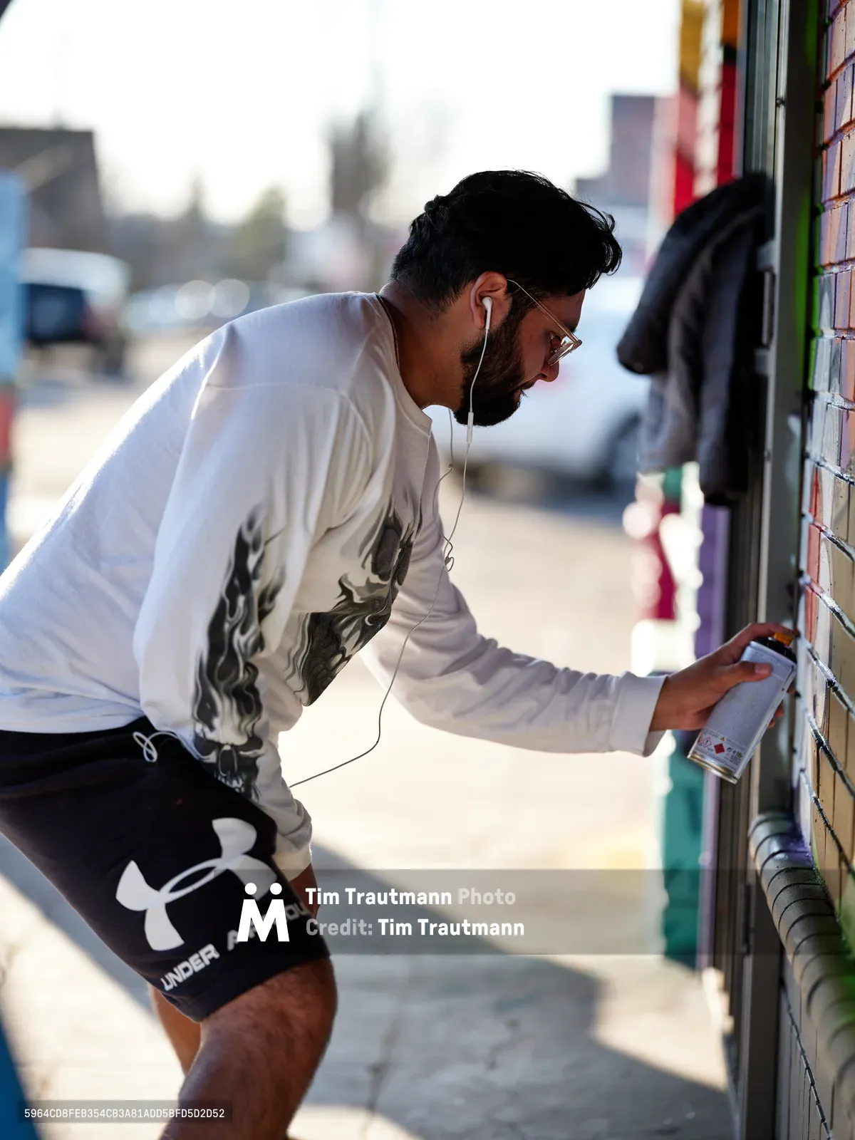 Muralist Heysus leans into his work along a vibrant wall at the intersection of Killingsworth and Northeast 15th Avenue in Portland's Alberta neighborhood. Bathed in warm afternoon light, the bearded artist wearing a white graphic long-sleeve shirt and black athletic shorts carefully selects from an array of spray paint cans, his white earbuds connecting him to his creative process. The urban streetscape behind him blurs into soft bokeh, while colorful building facades hint at the artistic energy that defines this gentrifying corner of Northeast Portland.