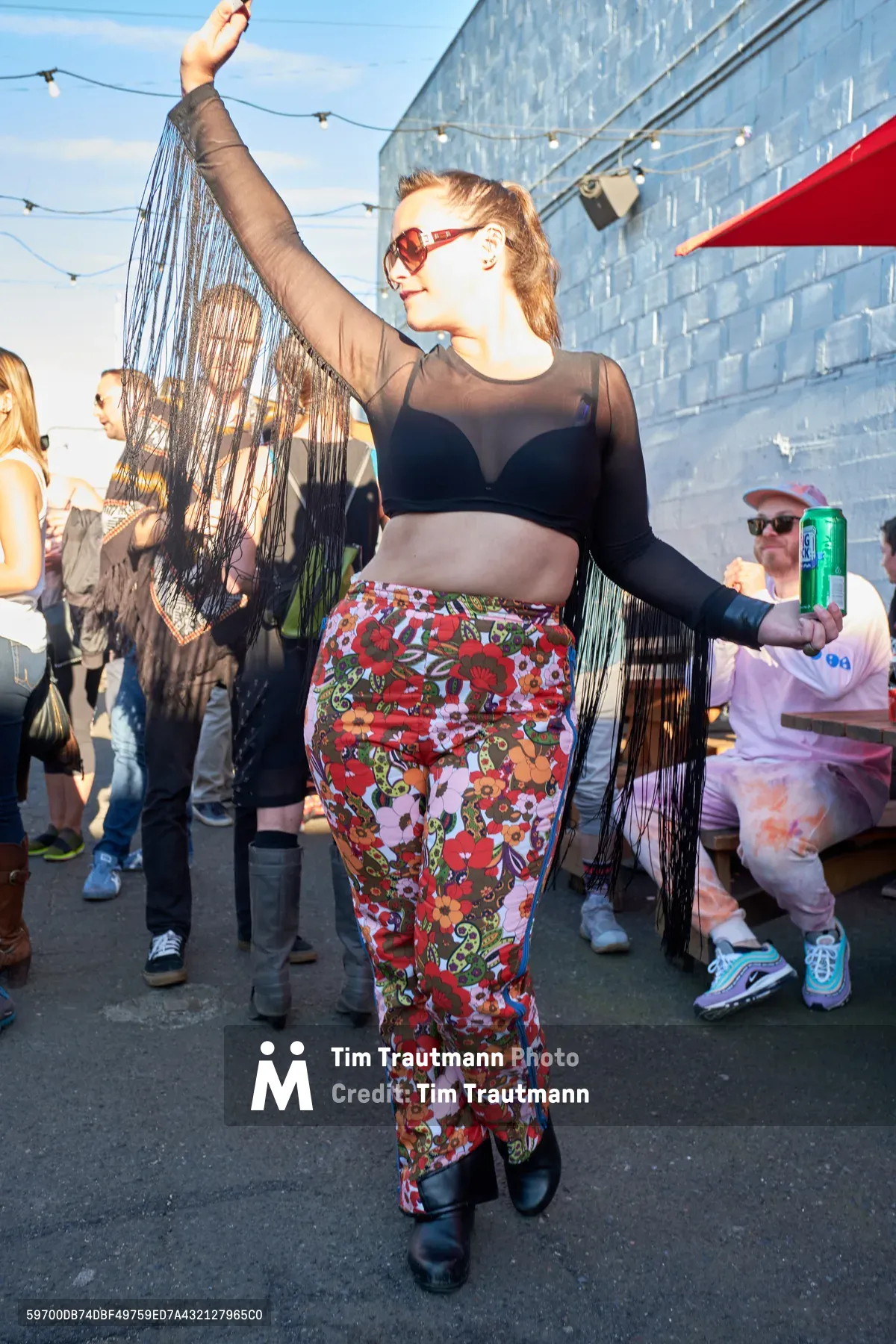 A young woman with sheer mesh sleeves and vintage floral pants moves ecstatically against Portland's blue brick architecture, her arms raised in pure celebration. The late afternoon sun bathes the White Owl Social Club rooftop in warm golden light, while string bulbs overhead and a relaxed crowd create an intimate day party atmosphere. Her burgundy sunglasses catch the light as cascading fringe details swing with her movement, embodying the carefree spirit of summer's first outdoor dance gathering.