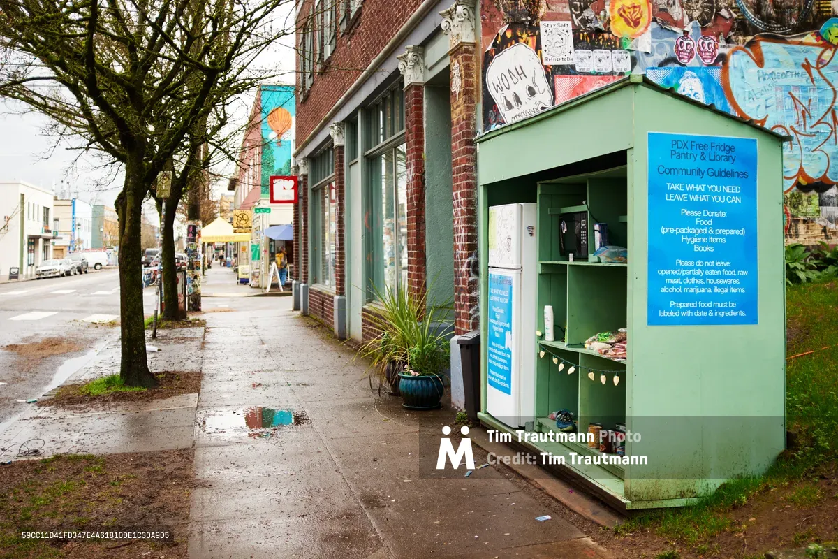 A mint green community refrigerator stands prominently on the sidewalk along Northeast 17th Avenue in Portland's Alberta Arts District, adorned with colorful murals and a bright blue informational panel. The weathered brick storefronts create a textured backdrop while bare winter trees cast intricate shadows across the damp pavement. This grassroots mutual aid station serves as a beacon of community solidarity, offering free food and supplies to neighbors in need.