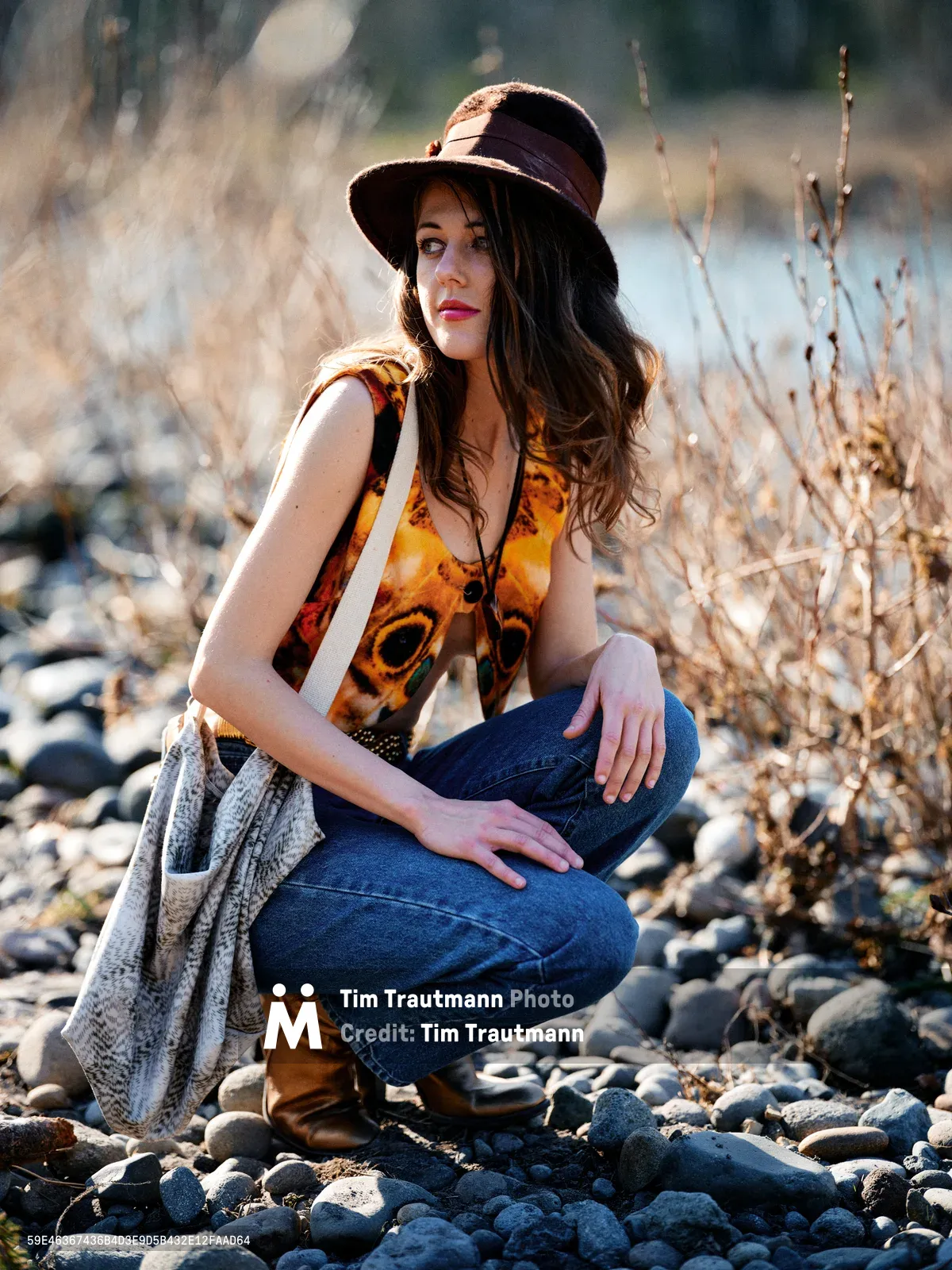 A young woman with flowing brunette hair sits contemplatively on a rocky riverbank along the Floodplain Trail in Multnomah County, Oregon. Bathed in warm golden hour light, she wears a distinctive amber-colored tank top with artistic face motifs, paired with blue jeans and a dark wide-brimmed hat. The natural setting features smooth river stones and dried winter grasses, with the Columbia River visible in the soft-focused background.