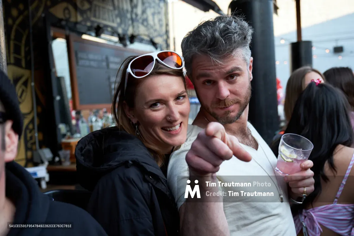 A jubilant couple shares an intimate moment at White Owl Social Club during Your Sunday Best, a popular summer day party in Portland, Oregon. The woman, wearing sunglasses and a black jacket, beams with joy as the bearded man in a cream henley playfully points toward the camera while holding a drink. The bustling venue atmosphere buzzes with conversation and laughter, captured in the warm, natural light filtering through the social club's windows.
