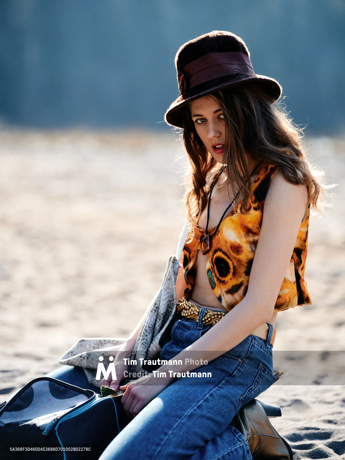 A young woman with flowing brown hair sits contemplatively on weathered driftwood along Oregon's Floodplain Trail in Multnomah County. Bathed in warm golden hour light, she wears a vintage brown felt hat with ribbon trim, a vibrant orange sunflower-patterned crop top, and classic blue jeans with a woven belt. Her leather messenger bag rests beside her as she gazes thoughtfully across the river bed terrain, embodying the spirit of Pacific Northwest exploration.