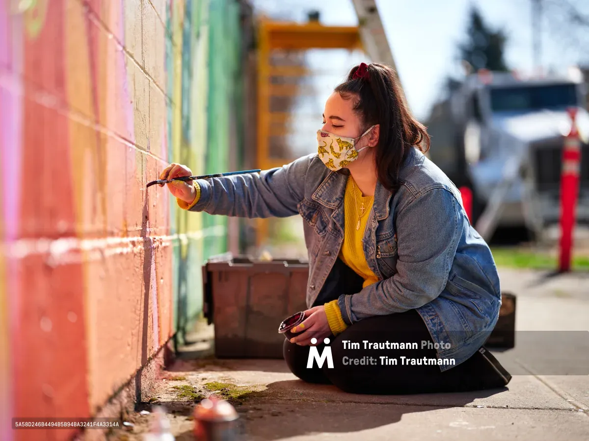 Multidisciplinary artist Vilatose sits cross-legged on the sidewalk, painting delicate details on her vibrant mural 'Jardín Cubana' depicting three female figures along Northeast Alberta Street in Portland's historic arts district. Wearing a patterned face mask and denim jacket over a golden yellow sweater, she extends her brush toward the rainbow-hued wall in warm afternoon light. The urban scene captures the intimate process of community art-making, with colorful buildings and parked vehicles softly blurred in the background.