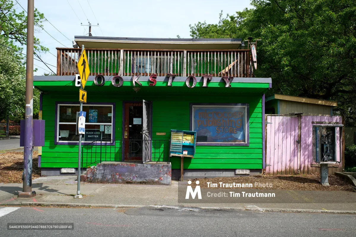 A vibrant emerald green independent bookstore stands along North Williams Avenue in Portland's historic Albina district. The weathered building features bold red lettering spelling 'BOOKSTORE' across its facade, with Microcosm Publishing's storefront window displaying community literature beneath a rustic wooden balcony. A public pay phone and lush Pacific Northwest foliage frame this cornerstone of Portland's literary and activist community.